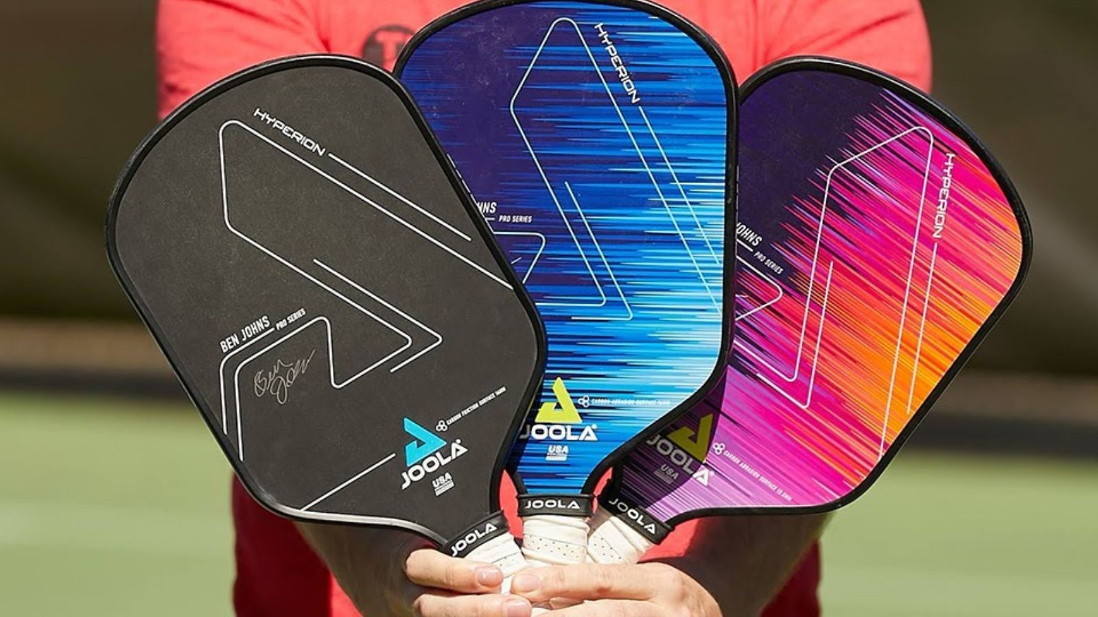Một người cầm ba cây vợt pickleball JOOLA Hyperion Ben Johns Pro Series với các thiết kế khác nhau: màu đen, xanh lam và tím-cam, đứng trước phông nền sân chơi.