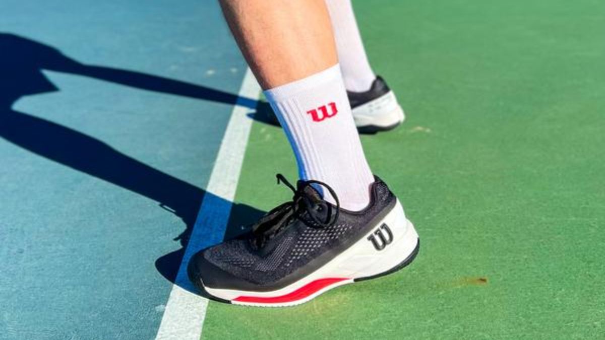 Người chơi tennis mang giày Wilson màu đen với chi tiết trắng và đỏ, kết hợp với tất trắng có logo Wilson, trên sân tennis ngoài trời.