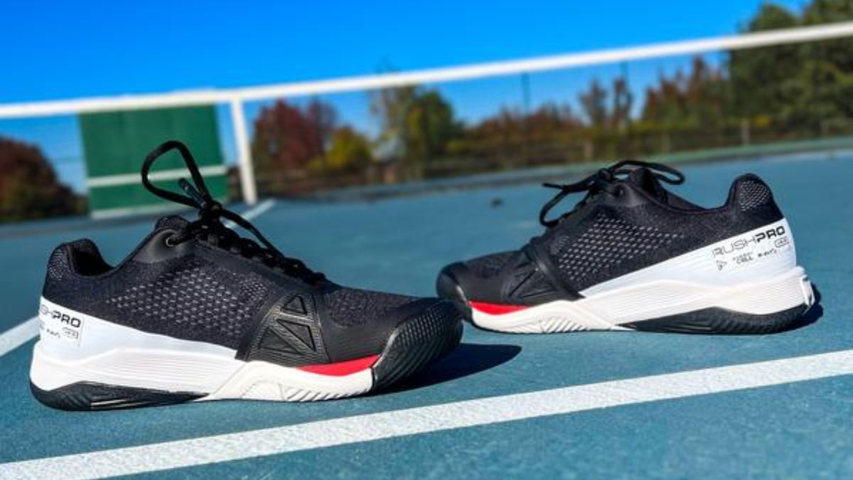 Một đôi giày tennis Wilson Rush Pro 4.0 trắng và đỏ, được đặt trên sân tennis ngoài trời dưới bầu trời trong xanh.